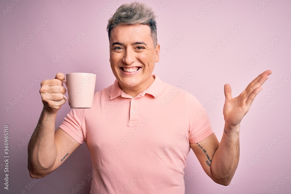 Young modern handsome man drinking a cup of hot coffee over isolated pink background very happy and excited, winner expression celebrating victory screaming with big smile and raised hands