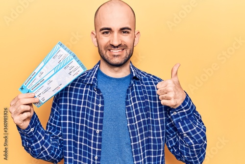 Young handsome bald man holding airline boarding pass smiling happy and positive, thumb up doing excellent and approval sign photo