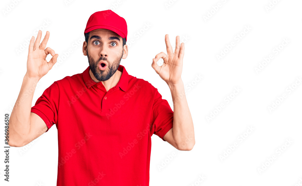 Young handsome man with beard wearing delivery uniform looking surprised and shocked doing ok approval symbol with fingers. crazy expression
