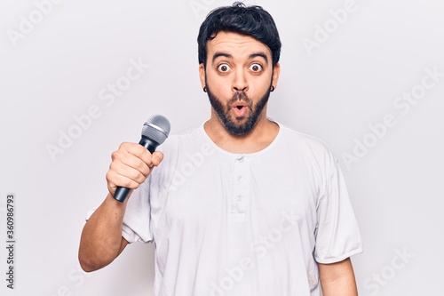 Young hispanic man singing song using microphone scared and amazed with open mouth for surprise, disbelief face