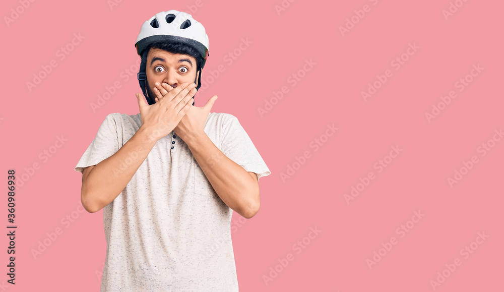 Young hispanic man wearing bike helmet shocked covering mouth with hands for mistake. secret concept.