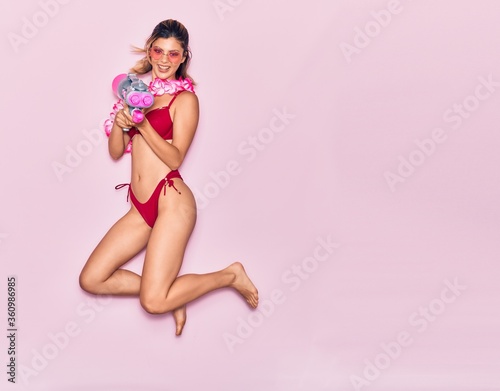 Fototapeta Naklejka Na Ścianę i Meble -  Young beautiful girl on vacation wearing bikini and hawaiian lei smiling happy. Jumping with smile on face holding water gun over isolated pink background