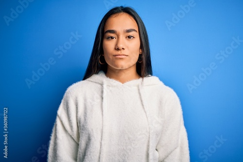 Young beautiful asian sportswoman wearing sweatshirt standing over isolated blue background with serious expression on face. Simple and natural looking at the camera.