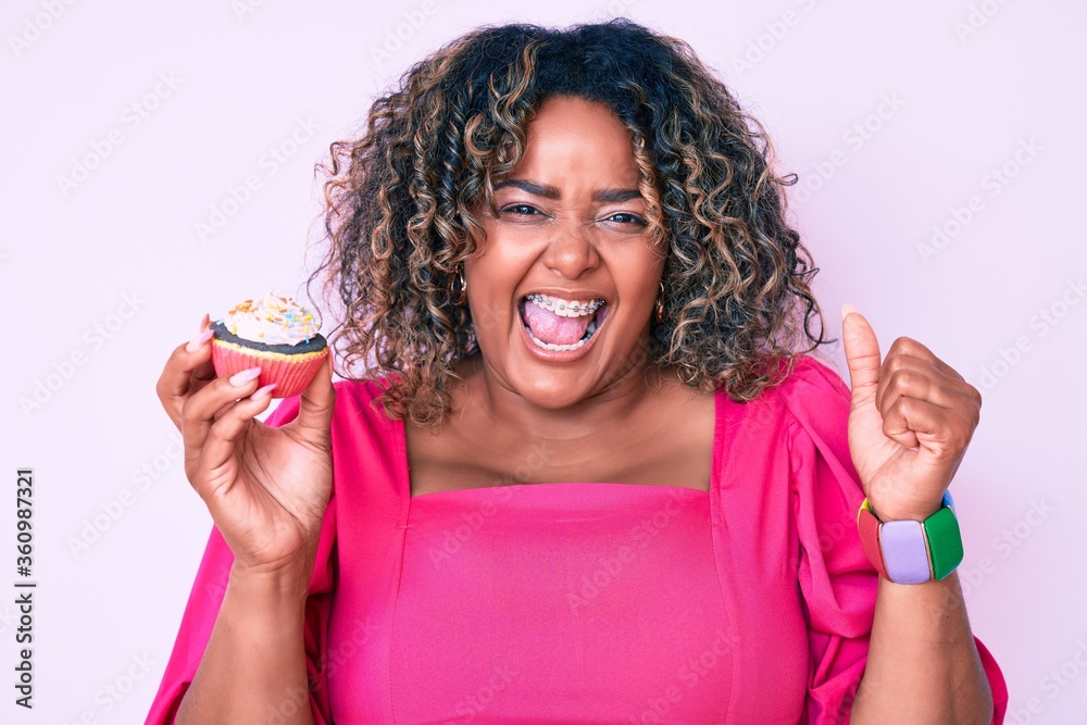 Young african american plus size woman holding cupcake screaming proud, celebrating victory and success very excited with raised arms Young african american plus size woman holding cupcake screaming proud, celebrating victory and success very excited with raised arms
