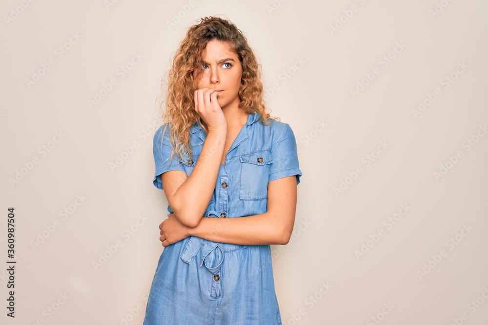 Young beautiful woman with blue eyes wearing casual denim dress over white background looking stressed and nervous with hands on mouth biting nails. Anxiety problem. Young beautiful woman with blue eyes wearing casual denim dress over white background looking stressed and nervous with hands on mouth biting nails. Anxiety problem.