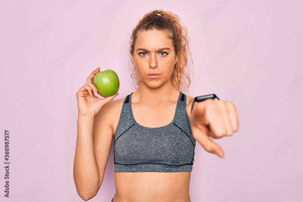 Young beautiful blonde sporty woman with blue eyes holding healthy green apple fruit pointing with finger to the camera and to you, hand sign, positive and confident gesture from the front Young beautiful blonde sporty woman with blue eyes holding healthy green apple fruit pointing with finger to the camera and to you, hand sign, positive and confident gesture from the front