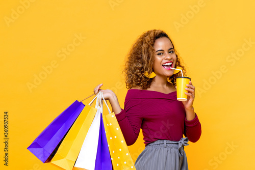 Portrait of cheerful young attractive African American woman holding shopping bags while drinking in isolated studio yellow background