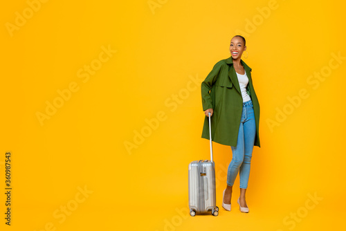 Full length portrait of travelling happy young African American tourist woman standing while holding luggage in isolated studio yellow background