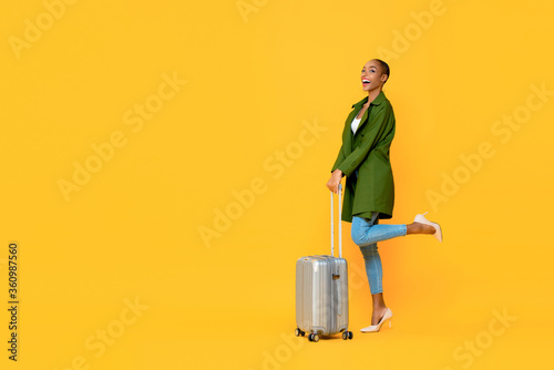 Full length portrait of excited young African American tourist woman holding luggage on one leg raised standing gesture ready to travel in isolated studio yellow background