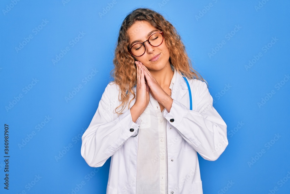 Young beautiful blonde doctor woman with blue eyes wearing coat and stethoscope sleeping tired dreaming and posing with hands together while smiling with closed eyes. Young beautiful blonde doctor woman with blue eyes wearing coat and stethoscope sleeping tired dreaming and posing with hands together while smiling with closed eyes.