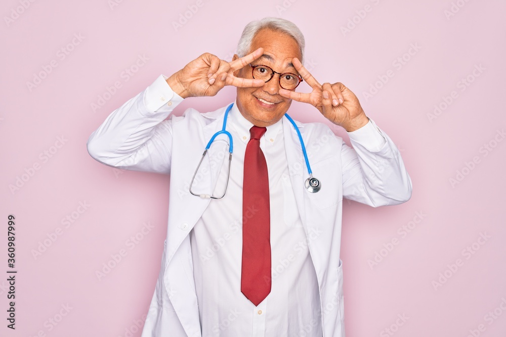 Middle age senior grey-haired doctor man wearing stethoscope and professional medical coat Doing peace symbol with fingers over face, smiling cheerful showing victory Middle age senior grey-haired doctor man wearing stethoscope and professional medical coat Doing peace symbol with fingers over face, smiling cheerful showing victory