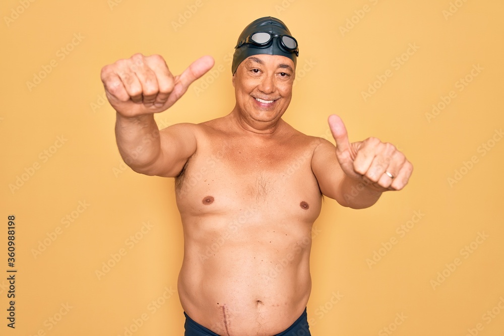 Middle age senior grey-haired swimmer man wearing swimsuit, cap and goggles approving doing positive gesture with hand, thumbs up smiling and happy for success. Winner gesture.