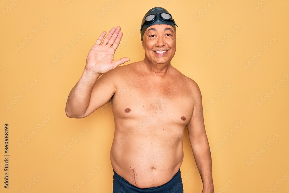 Middle age senior grey-haired swimmer man wearing swimsuit, cap and goggles Waiving saying hello happy and smiling, friendly welcome gesture