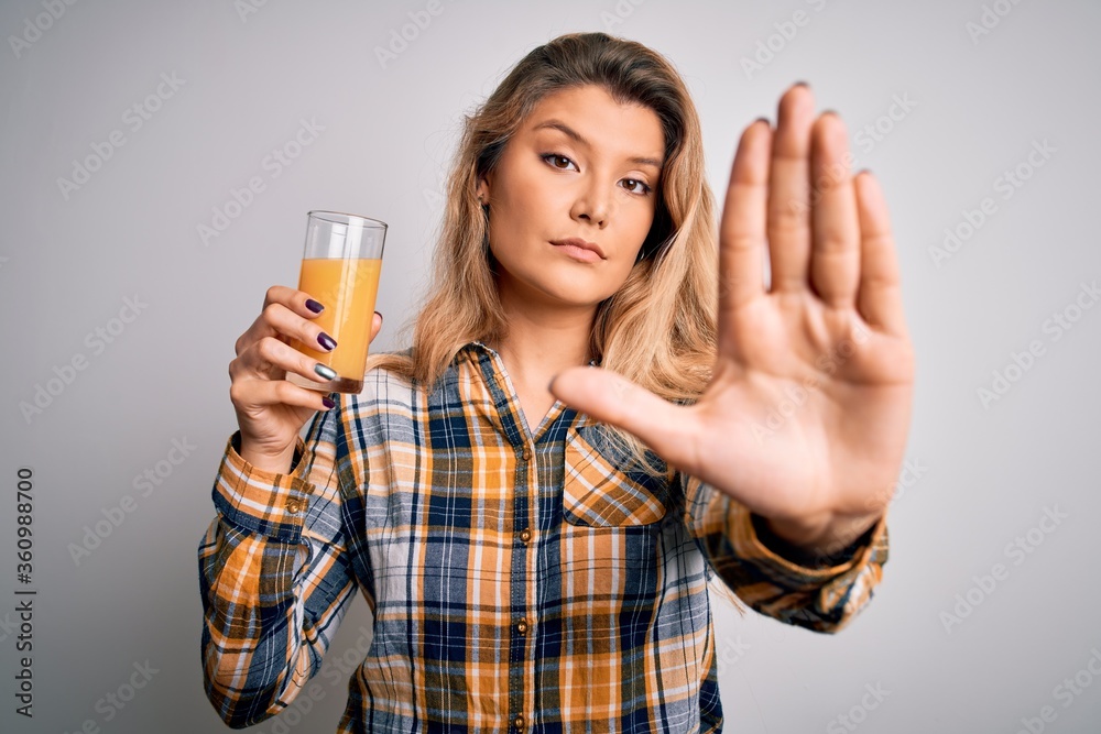 Young beautiful blonde woman drinking glass of healthy orange juice over white background with open hand doing stop sign with serious and confident expression, defense gesture Young beautiful blonde woman drinking glass of healthy orange juice over white background with open hand doing stop sign with serious and confident expression, defense gesture