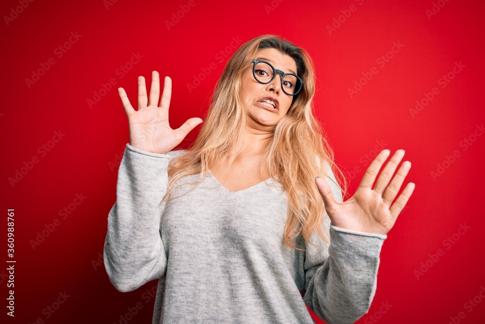 Young beautiful blonde woman wearing sweater and glasses over isolated red background afraid and terrified with fear expression stop gesture with hands, shouting in shock. Panic concept. Young beautiful blonde woman wearing sweater and glasses over isolated red background afraid and terrified with fear expression stop gesture with hands, shouting in shock. Panic concept.