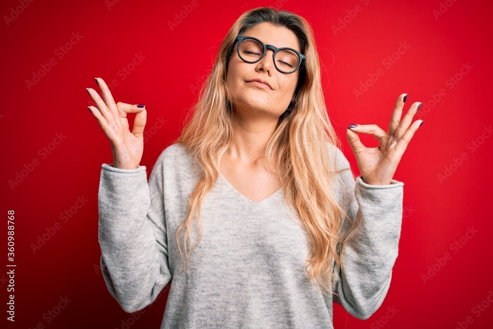 Young beautiful blonde woman wearing sweater and glasses over isolated red background relax and smiling with eyes closed doing meditation gesture with fingers. Yoga concept.