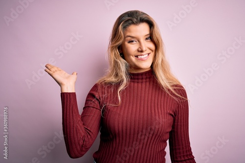 Young beautiful blonde woman wearing casual sweater over isolated pink background smiling cheerful presenting and pointing with palm of hand looking at the camera.