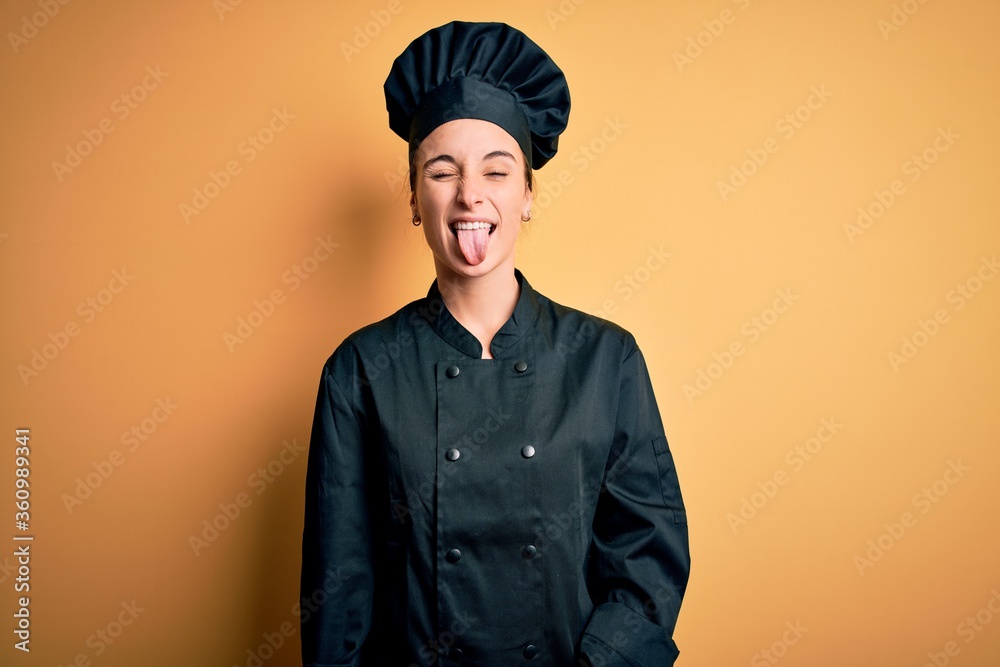 Young beautiful chef woman wearing cooker uniform and hat standing over yellow background sticking tongue out happy with funny expression. Emotion concept. Young beautiful chef woman wearing cooker uniform and hat standing over yellow background sticking tongue out happy with funny expression. Emotion concept.