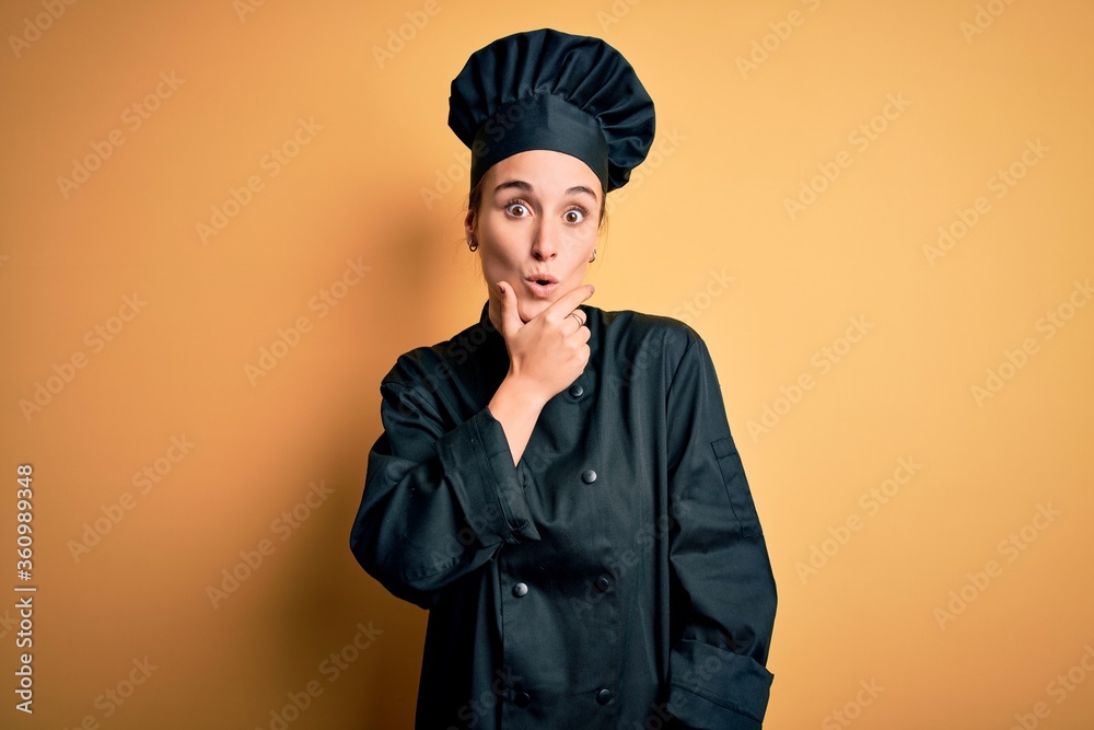 Young beautiful chef woman wearing cooker uniform and hat standing over yellow background Looking fascinated with disbelief, surprise and amazed expression with hands on chin Young beautiful chef woman wearing cooker uniform and hat standing over yellow background Looking fascinated with disbelief, surprise and amazed expression with hands on chin
