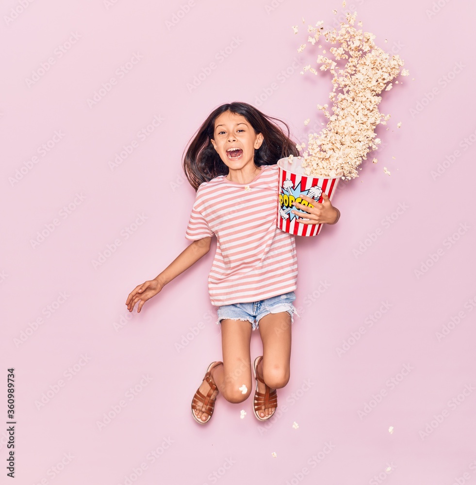 Adorable hispanic child girl surprised with open mouth holding bucket of popcorn jumping over isolated pink background Adorable hispanic child girl surprised with open mouth holding bucket of popcorn jumping over isolated pink background