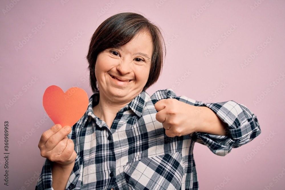 Young down syndrome romantic woman holding red heart paper shape over pink background screaming proud and celebrating victory and success very excited, cheering emotion Young down syndrome romantic woman holding red heart paper shape over pink background screaming proud and celebrating victory and success very excited, cheering emotion