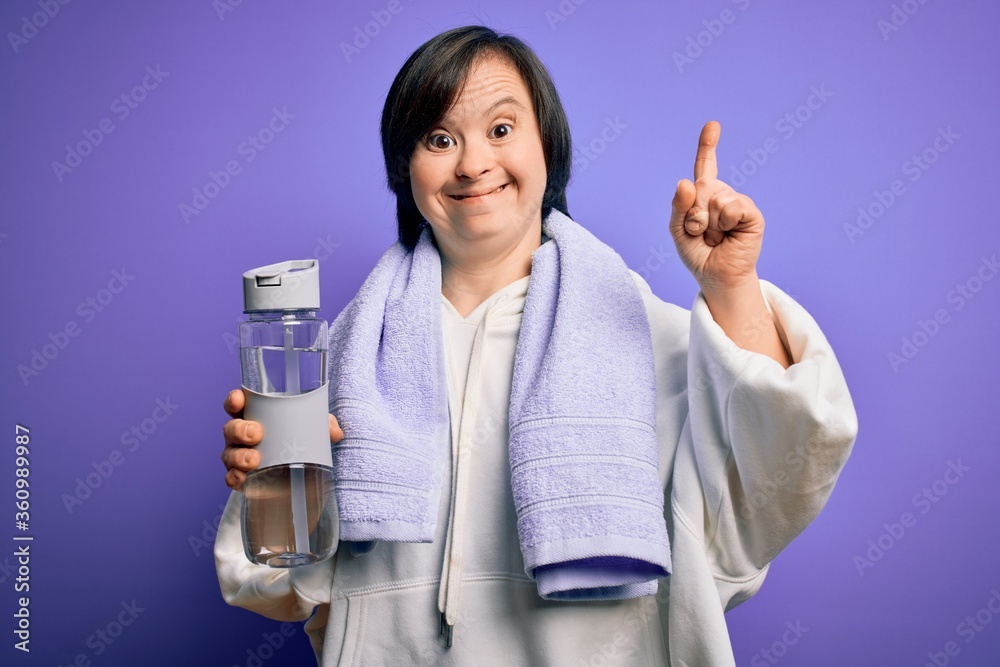 Young down syndrome fitness woman training at gym holding water bottle and sport towel surprised with an idea or question pointing finger with happy face, number one Young down syndrome fitness woman training at gym holding water bottle and sport towel surprised with an idea or question pointing finger with happy face, number one