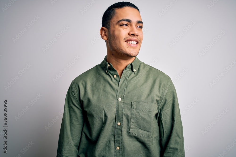 Young brazilian man wearing casual shirt standing over isolated white background looking away to side with smile on face, natural expression. Laughing confident.