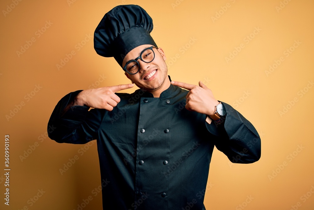 Young brazilian chef man wearing cooker uniform and hat over isolated yellow background smiling cheerful showing and pointing with fingers teeth and mouth. Dental health concept. Young brazilian chef man wearing cooker uniform and hat over isolated yellow background smiling cheerful showing and pointing with fingers teeth and mouth. Dental health concept.