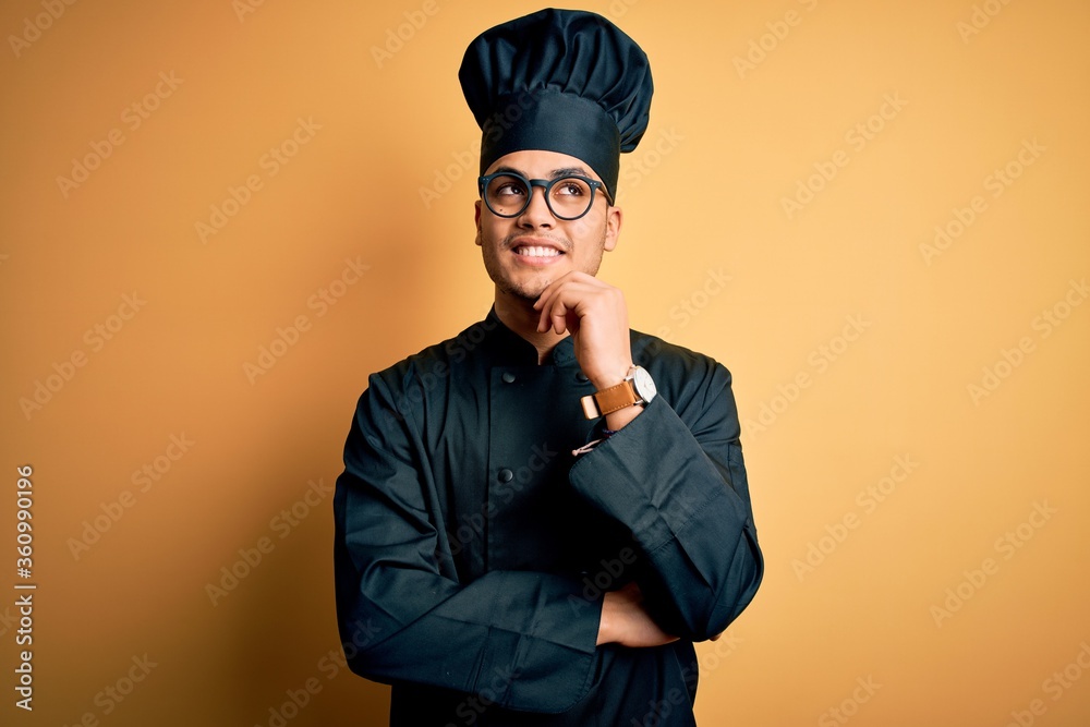 Young brazilian chef man wearing cooker uniform and hat over isolated yellow background with hand on chin thinking about question, pensive expression. Smiling and thoughtful face. Doubt concept. Young brazilian chef man wearing cooker uniform and hat over isolated yellow background with hand on chin thinking about question, pensive expression. Smiling and thoughtful face. Doubt concept.