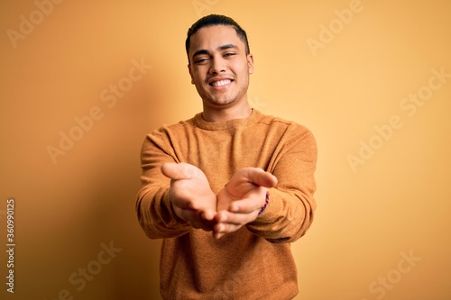 Young brazilian man wearing casual sweater standing over isolated yellow background Smiling with hands palms together receiving or giving gesture. Hold and protection