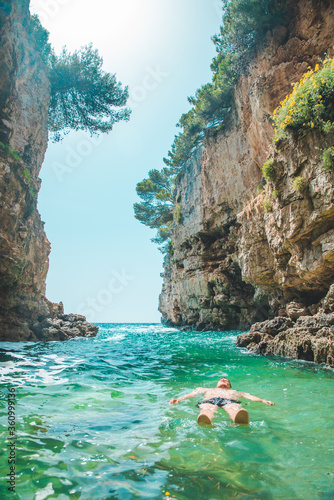 young man swimming on back in sea at bay between cliffs