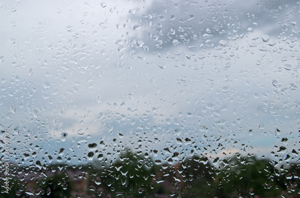 Abstract pattern consisting of randomly arranged raindrops hanging on window pane after recent rain with background of blurred foliage of tree tops, steel-blue sky with light and dark bizarre clouds Abstract pattern consisting of randomly arranged raindrops hanging on window pane after recent rain with background of blurred foliage of tree tops, steel-blue sky with light and dark bizarre clouds