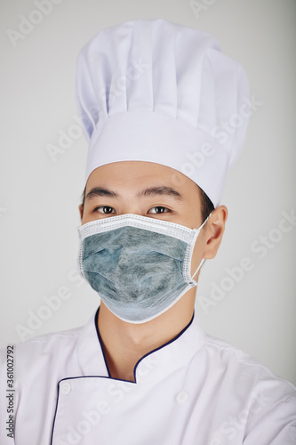 Portrait of young serious Asian restaurant chef in hat and medical mask looking at camera