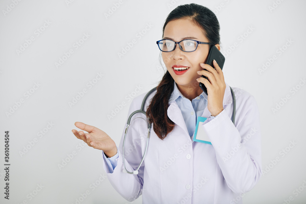 Excited young Asian general practitioner in glasses talking on phone with colleague or patient Excited young Asian general practitioner in glasses talking on phone with colleague or patient