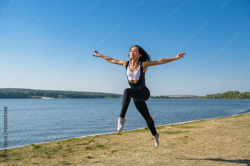 Obraz premium Smiling young female gymnast is jumping in split outdoors near the lake. Healthy lifestyle