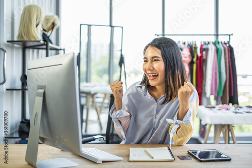 Young Asian woman business owner sitting at a desk in a clothing shop looking at a computer monitor with a happy cheerful expression on her face.
