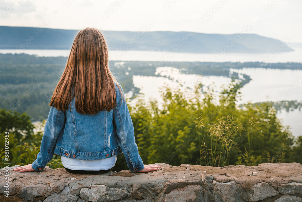 A little girl child in a denim jacket and with long hair sits on the edge of a cliff holding her hat with her hands and looks at the amazing natural picture with mountains and a river, the concept of 