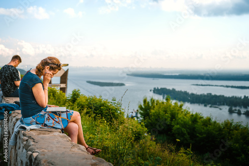 A beautiful stranger a woman in a skirt sits on a rock and reads a book against the background of a panorama of mountains and a river