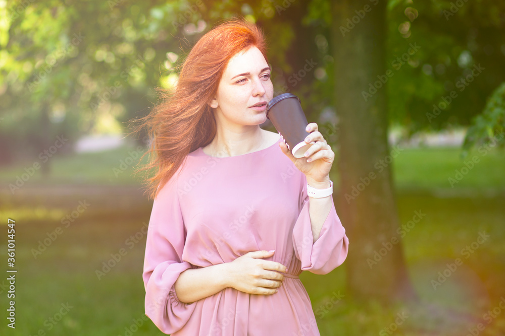 Young, red-haired woman walks on a summer day in the park, outdoors. Drinking coffee, to go