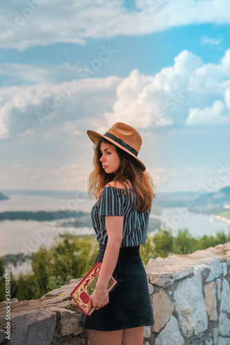 Woman traveler with long hair holding hat and looking at amazing mountains and river, wanderlust travel concept, space for text, atmospheric epic moment © KseniaJoyg