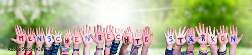 Children Hands Building Colorful German Word Menschenrechte Staerken Means Strengthen Human Rights. Sunny Green Grass Meadow As Background