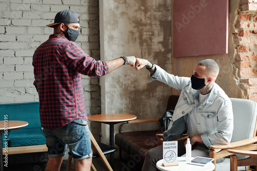 Young man in facial mask and ball cap making fist bump with friend while greeting him in lobby