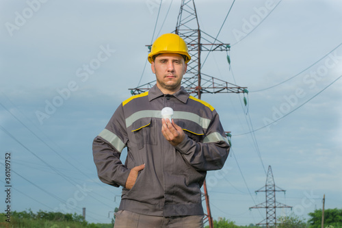 The power engineer holds an economic light bulb with one hand. In the background, a high-voltage support. Worker in a protective helmet. Industry. Energy. Electricity.