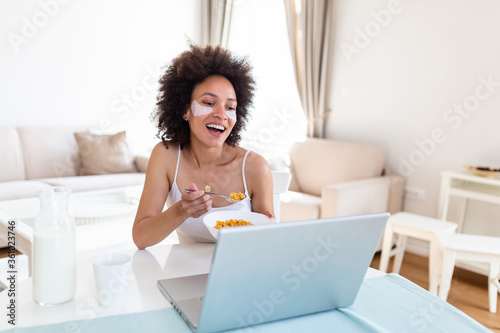 Image of happy young amazing woman sitting indoors at the table with laptop holding corn flakes. Looking at laptop computer and talking to her friends via video call.