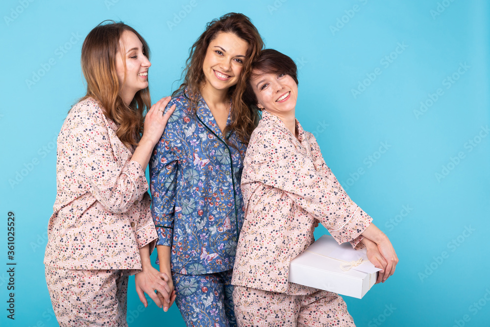 Portrait of three beautiful young girls wearing colorful pyjamas having fun during sleepover isolated over blue background. Pajama party and hen-party concept