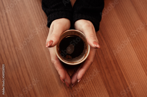 Top close up view of female hands with beautiful manicure hold eco friendly natural disposable cup of hot dark coffee on the wooden table in coffee house. Image of ecological dish with copy space. 