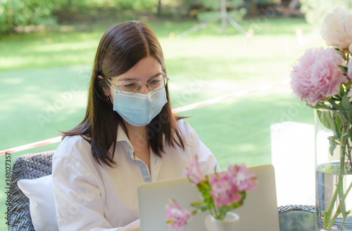 a middle-aged brunette girl in a protective face mask works at a laptop on the summer veranda of a cafe, a business concept after the coronavirus pandemic photo