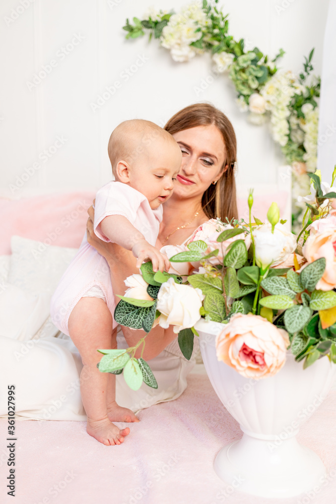 a young beautiful mother holds her daughter a girl of 6 months on a white bed and looks at flowers, a place for text a young beautiful mother holds her daughter a girl of 6 months on a white bed and looks at flowers, a place for text