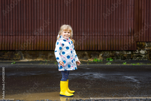 Little toddler girl wearing yellow rain boots, running and walking during sleet on rainy cloudy day. Cute happy child in colorful clothes jumping into puddle, splashing with water, outdoor activity