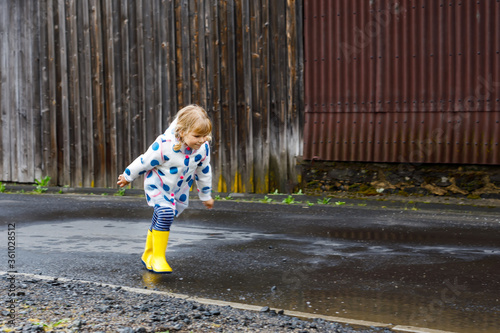 Little toddler girl wearing yellow rain boots, running and walking during sleet on rainy cloudy day. Cute happy child in colorful clothes jumping into puddle, splashing with water, outdoor activity